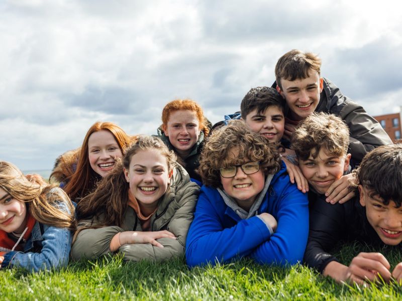Front view of a large group of teenage friends wearing casual clothing on a summer day in Whitley Bay, Northeastern England. They are gathered on the grass together, lying on their fronts smiling and looking at the camera.